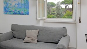 Un canapé gris avec un coussin, une fenêtre donnant sur un paysage verdoyant et un tableau accroché au mur.