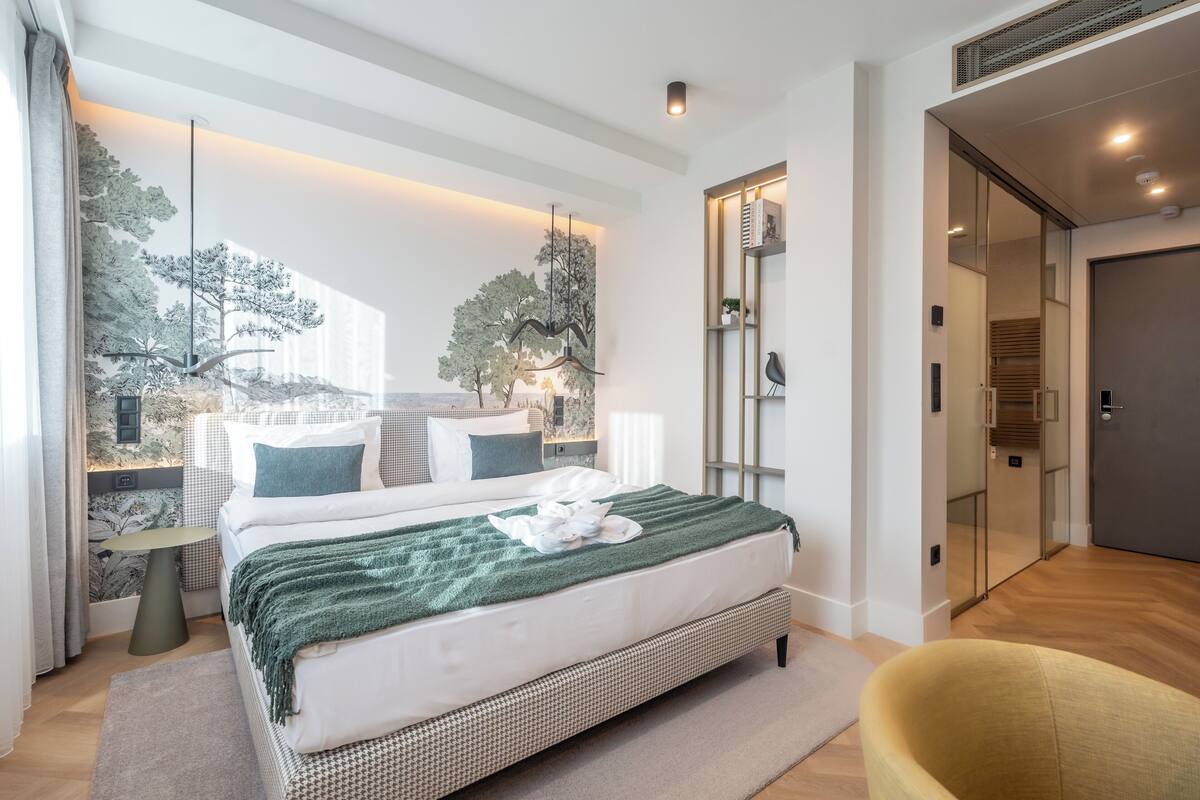 A modern bedroom with a large bed, a green throw blanket, and a view of trees through a window.