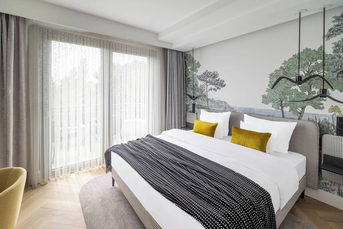 A modern hotel room with a large bed, a view of the outdoors through sheer curtains, and a mural of trees on the wall.