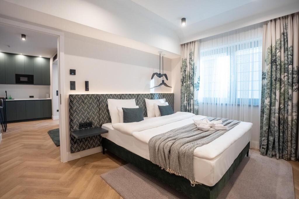 A modern bedroom with a large bed, a bedside table, a built-in bench, and a view of a kitchen area.