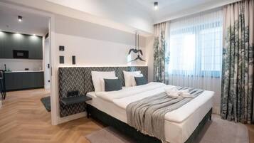 A modern bedroom with a large bed, a bedside table, a built-in bench, and a view of a kitchen area.