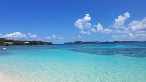 Una playa con agua turquesa cristalina, una orilla arenosa e islas lejanas bajo un cielo azul con nubes.