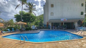 Una piscina con solárium de baldosas, un restaurante llamado "Sadi's" con terraza, y un edificio con techo de paja.