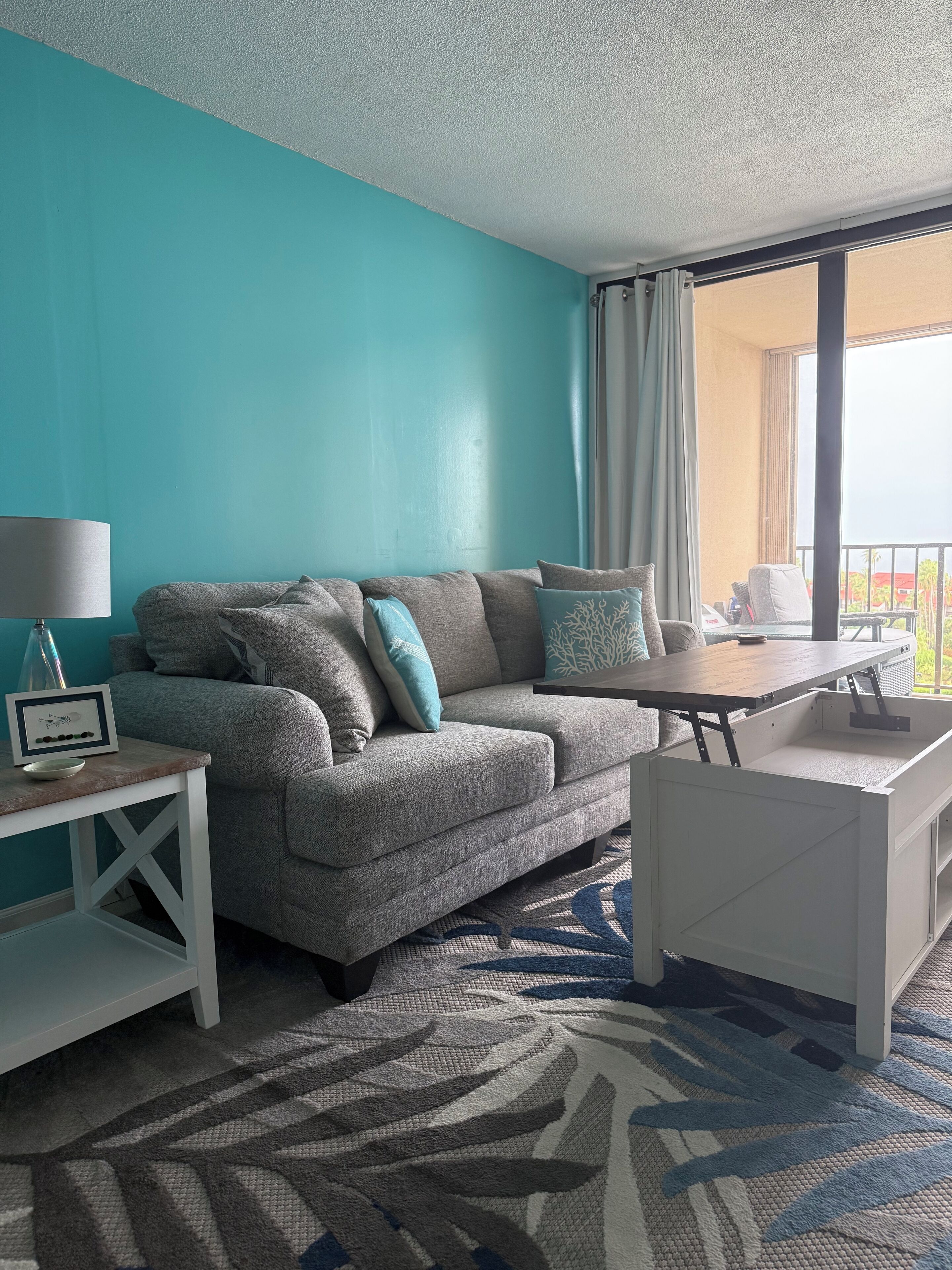 Un salon moderne avec un mur de couleur turquoise, un canapé gris agrémenté de coussins décoratifs, une petite table d’appoint blanche avec une lampe, et une grande fenêtre avec des rideaux.