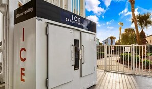 A 24-hour vending machine labeled "ICE" with a "24 hour vending" sign, located outside a building with a white fence and palm trees in the background. - Surf's Up Beachfront Resort Studio | Pools + Beach Access (Gulf Shores)