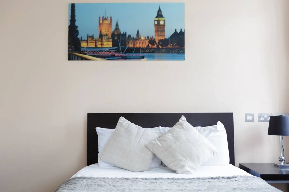 A hotel room with a bed featuring white bedding and two pillows, a nightstand with a lamp, and a wall-mounted painting of a cityscape.