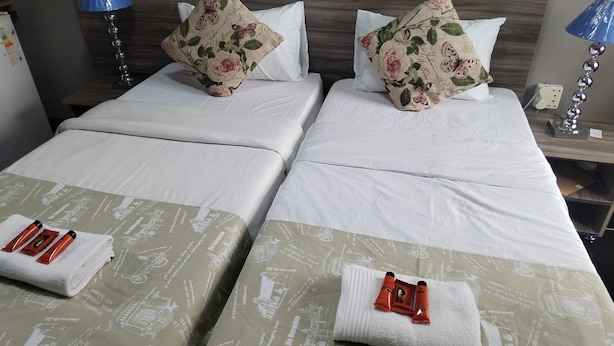 A hotel room with two beds, each with a floral pillow and a white bedsheet. There are two bedside lamps, one blue and one silver. - Room-home of Peace and Tranquility (Pretoria)