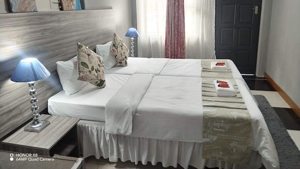 A hotel room with a large bed, two bedside lamps, a window with sheer curtains, and a door. - Room-home of Peace and Tranquility (Pretoria)