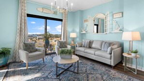 A living room with a sofa, two armchairs, a coffee table, and a chandelier. There is a large window with a view of the city.