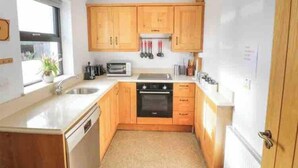 A compact kitchen with wooden cabinets, a dishwasher, a stove, and a window providing natural light. - Rose Cottage in Ennistymon - Near Cliffs of Moher (Magherareagh)
