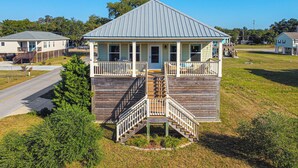 A two-story wooden house with a porch and a staircase, surrounded by a grassy area and a road.