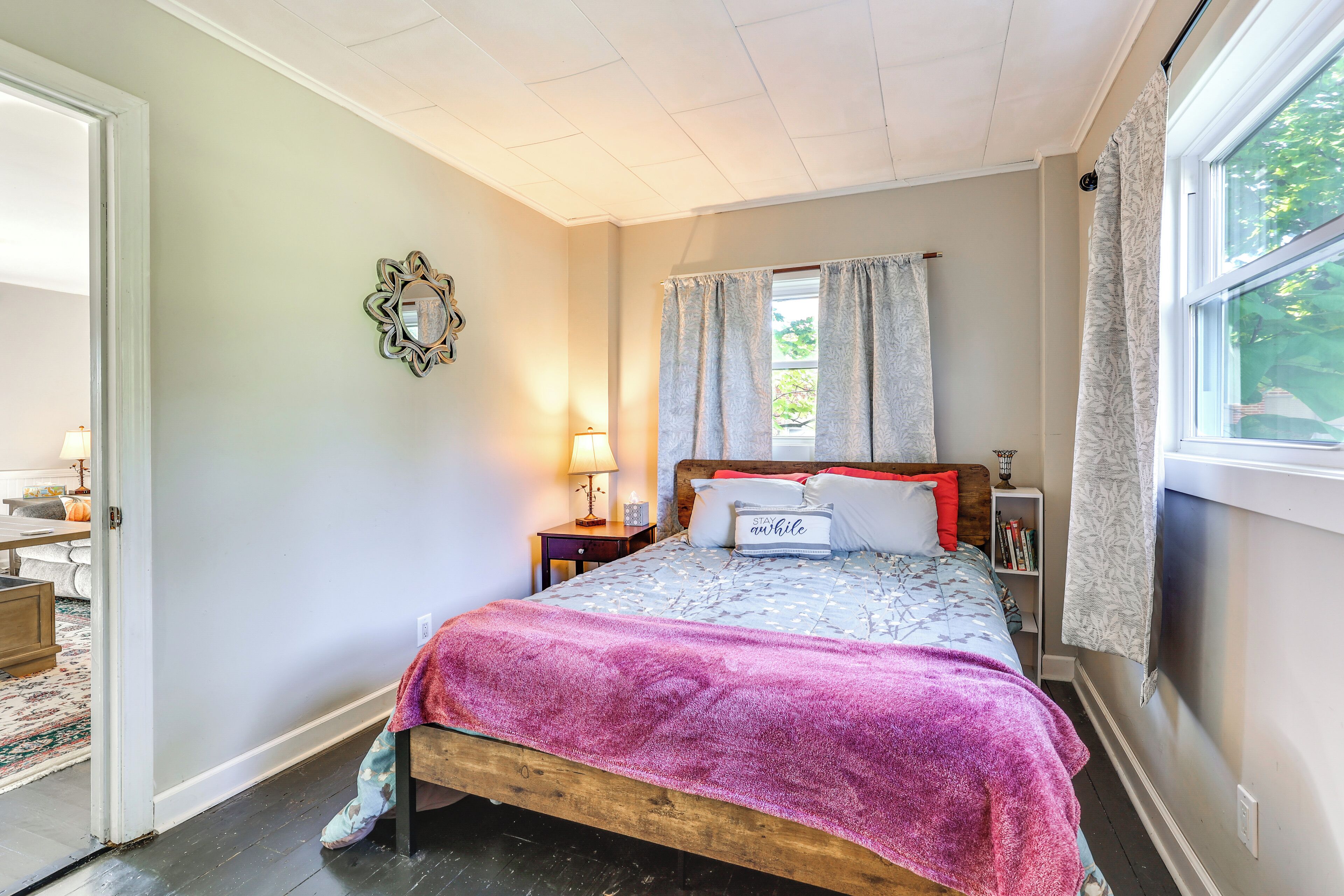 Een slaapkamer met een houten bed, een nachtkastje met een lamp, een raam met gordijnen en een deur die naar een andere kamer leidt.
