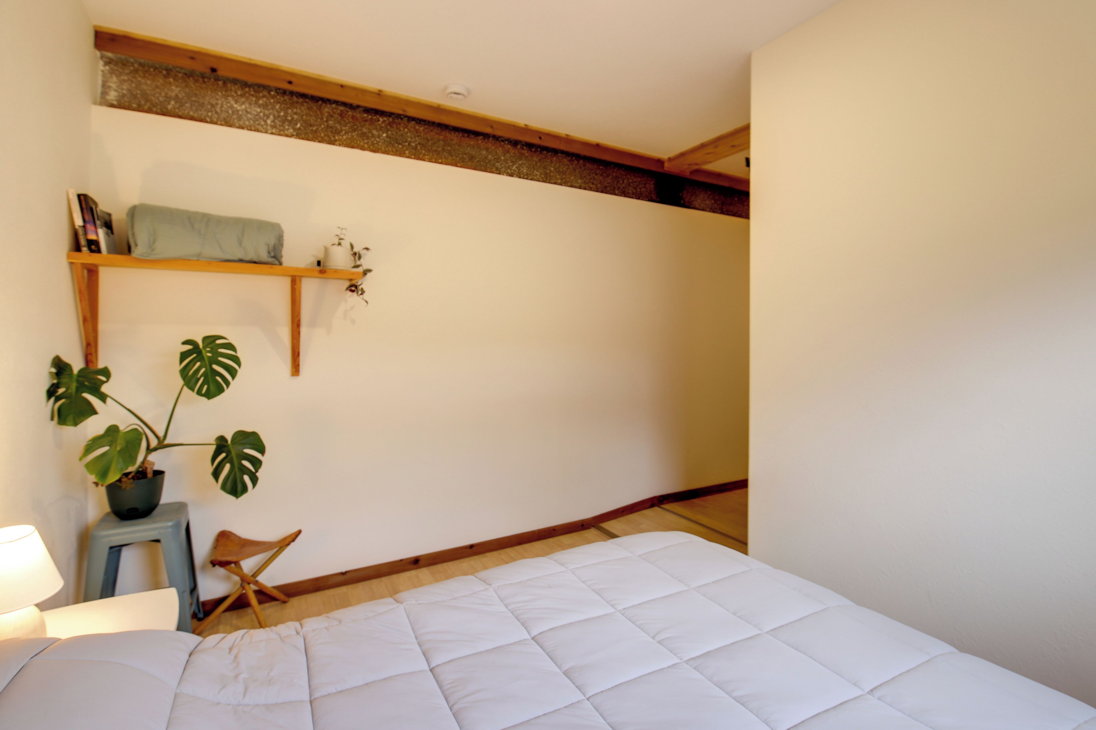 Un dormitorio con cama, un taburete, una silla, una estantería con libros y una planta, y techo con vigas de madera.