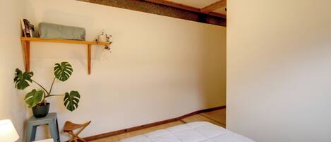 A bedroom with a bed, a small stool, a chair, a shelf with books and a plant, and a wooden beam ceiling.