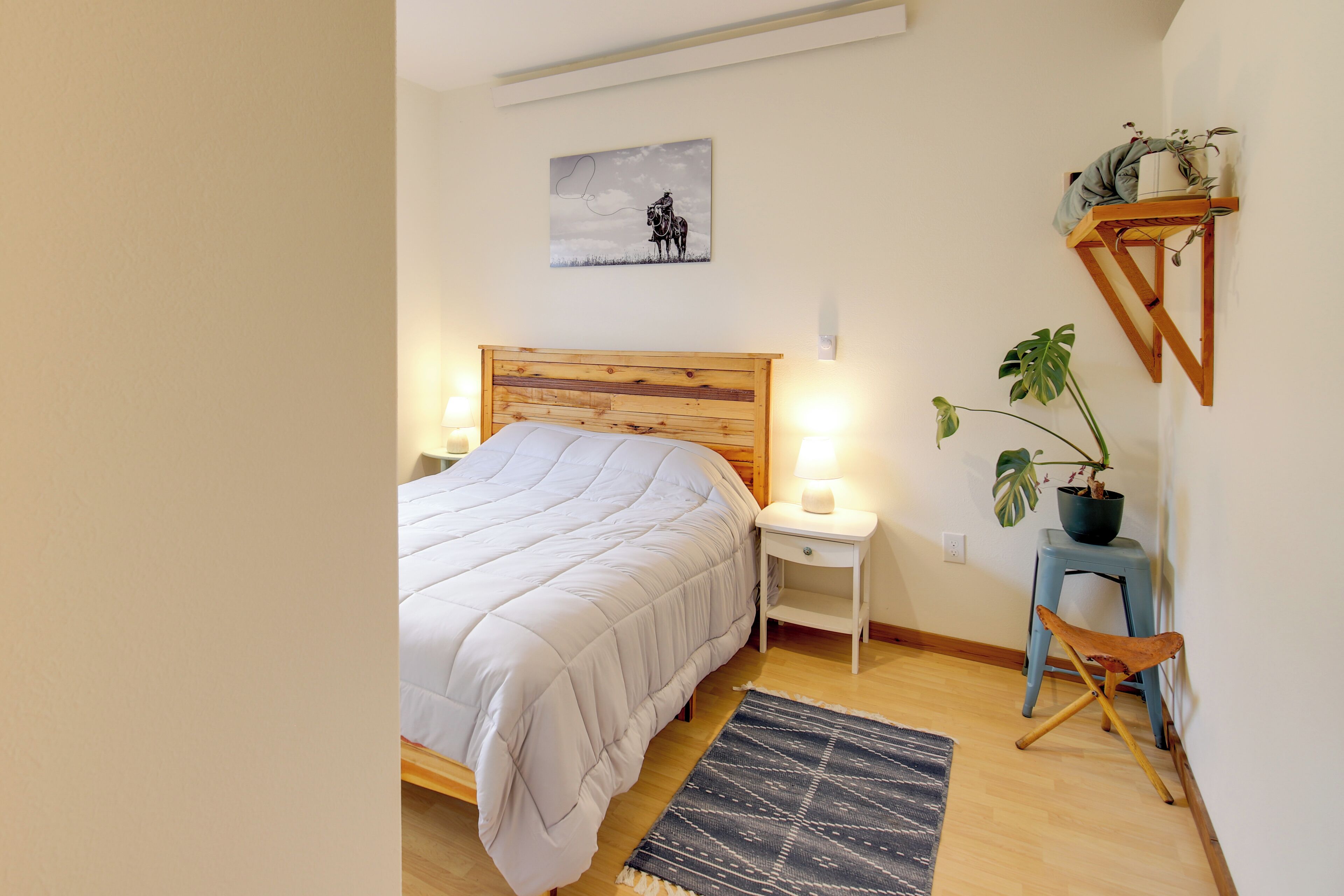 Een slaapkamer met een bed met houten hoofdbord, nachtkastjes met lampen, een potplant en een wandplank met decoratieve objecten.