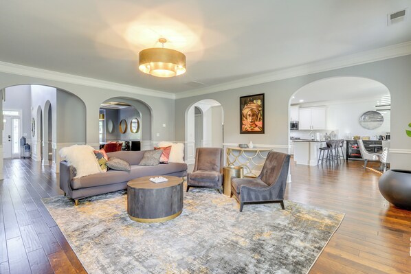 A spacious living room with a grey sofa, two armchairs, a coffee table, and a rug. There is a kitchen in the background with white cabinets and a dining area.