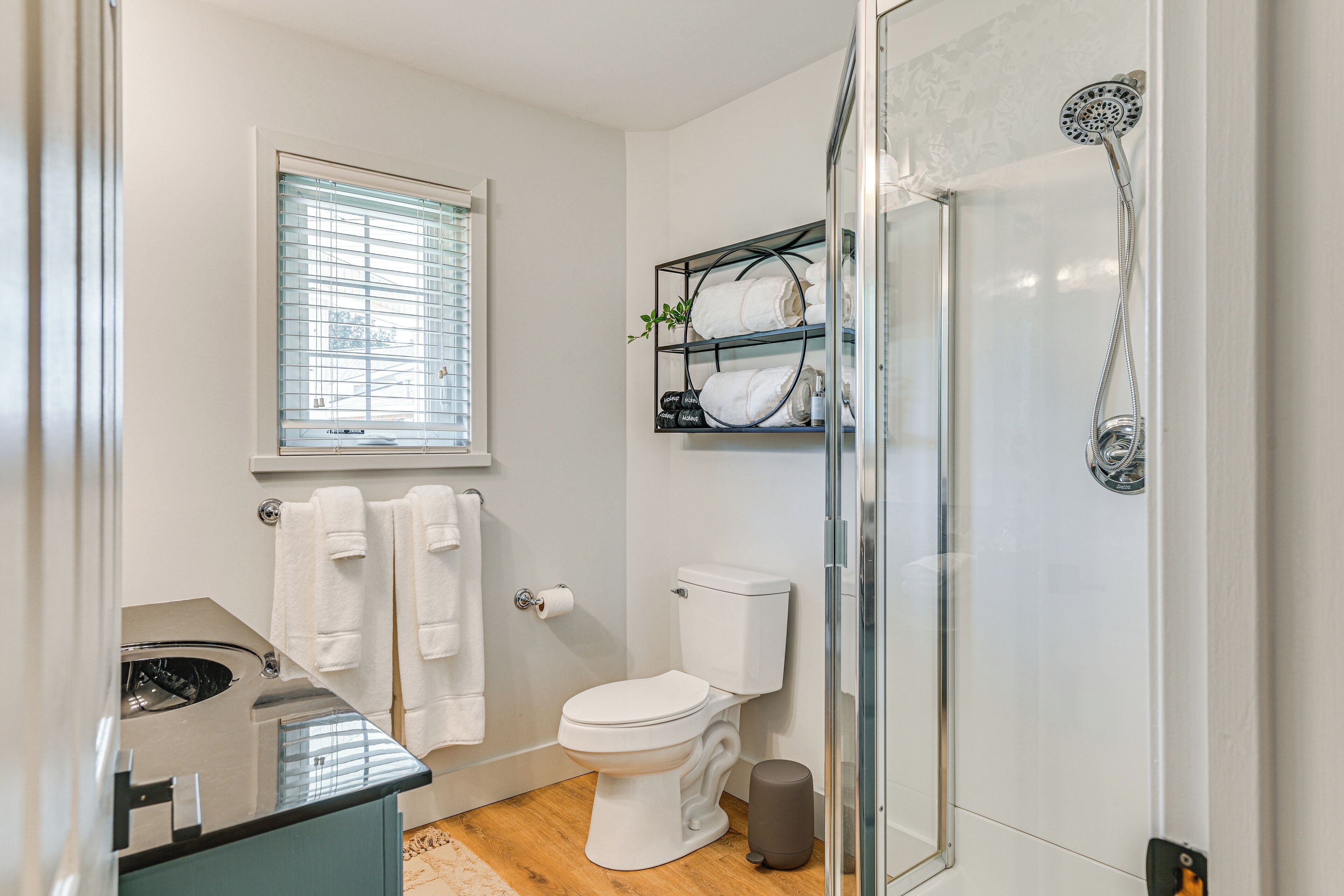 A modern bathroom with a toilet, a glass shower enclosure, a towel rack with towels, and a window with blinds.