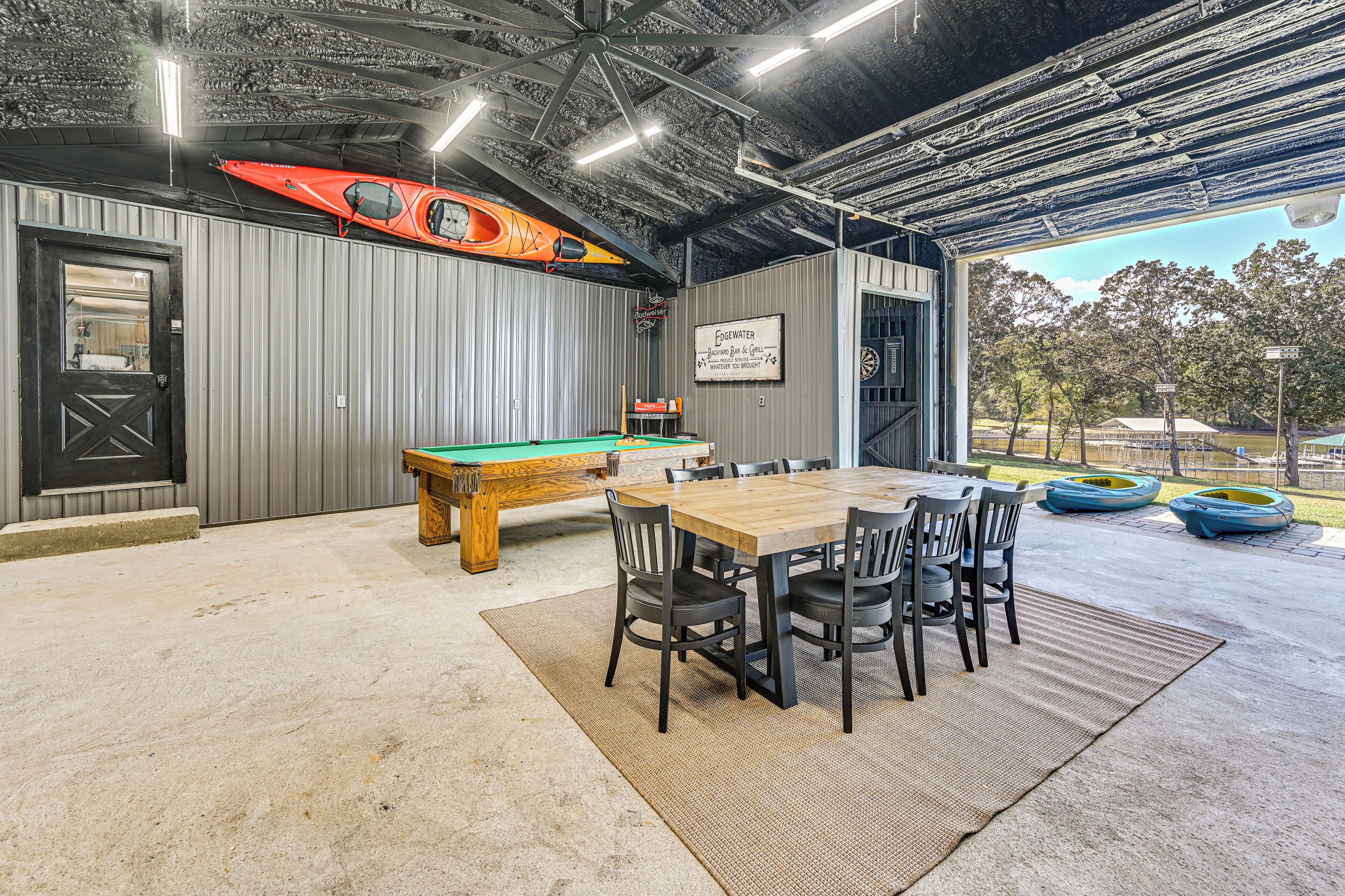 Un espace extérieur abrité comprenant une table de billard, une table en bois avec des chaises et des kayaks appuyés contre le mur.