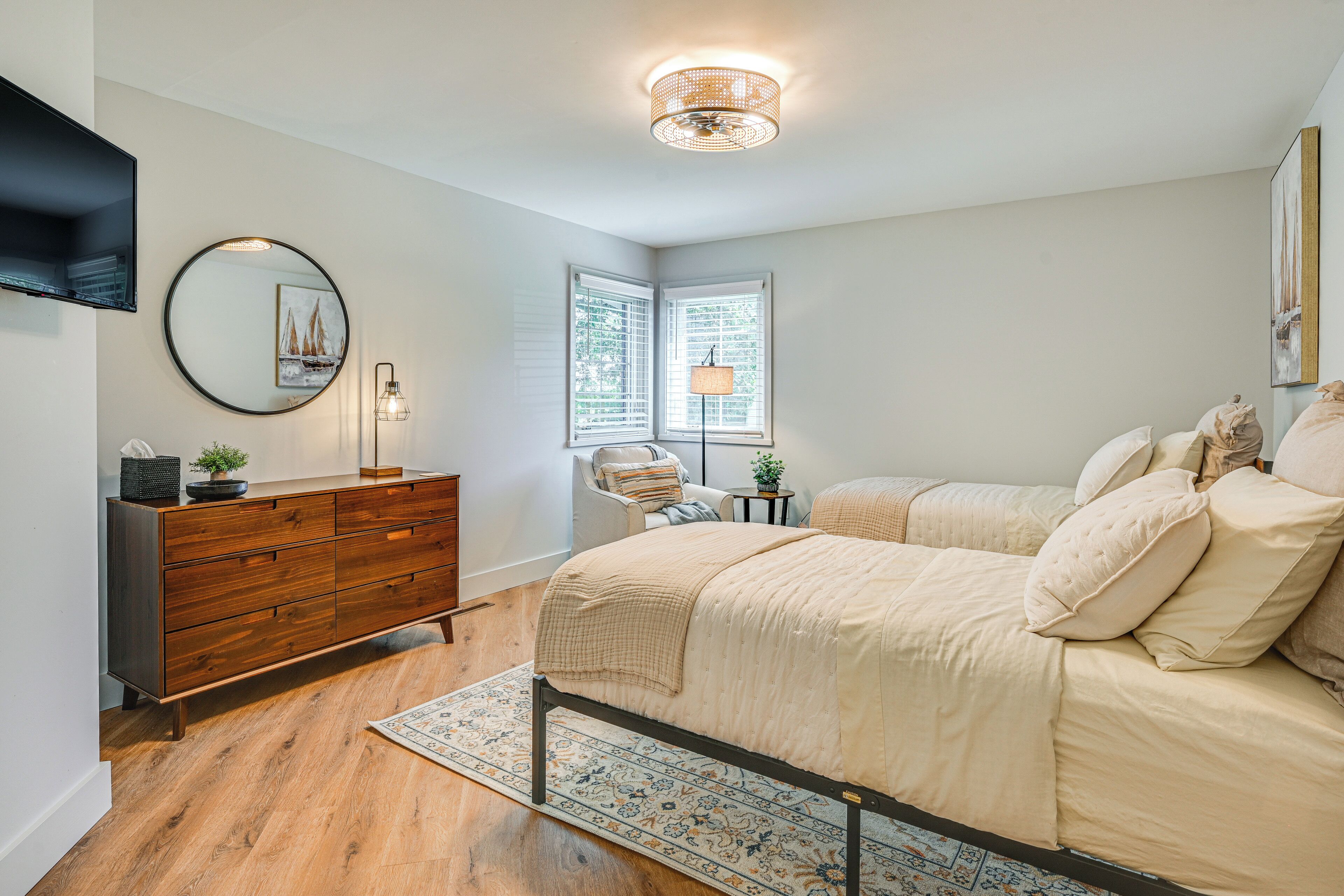 A bedroom with a wooden dresser, a round mirror, a bed with beige bedding, a floor lamp, and a window with blinds.