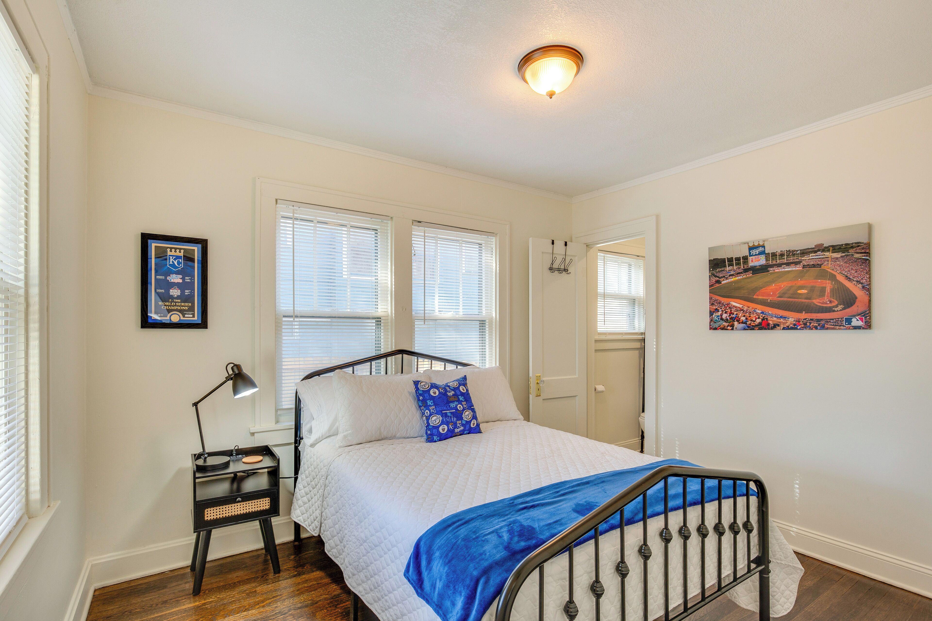 Quarto com cama, mesa de cabeceira, luminária, janela com persianas, porta e um quadro na parede.