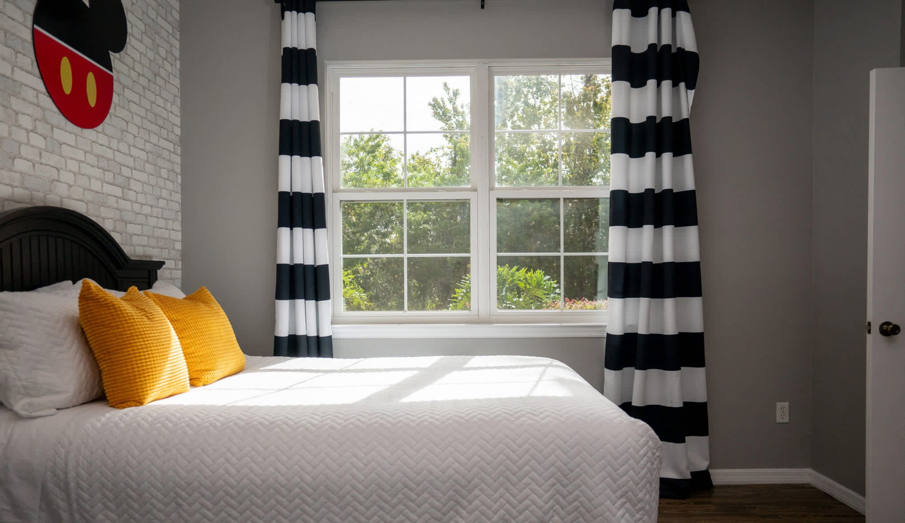 A bedroom with a bed, a window with black and white striped curtains, and a Mickey Mouse wall decoration.