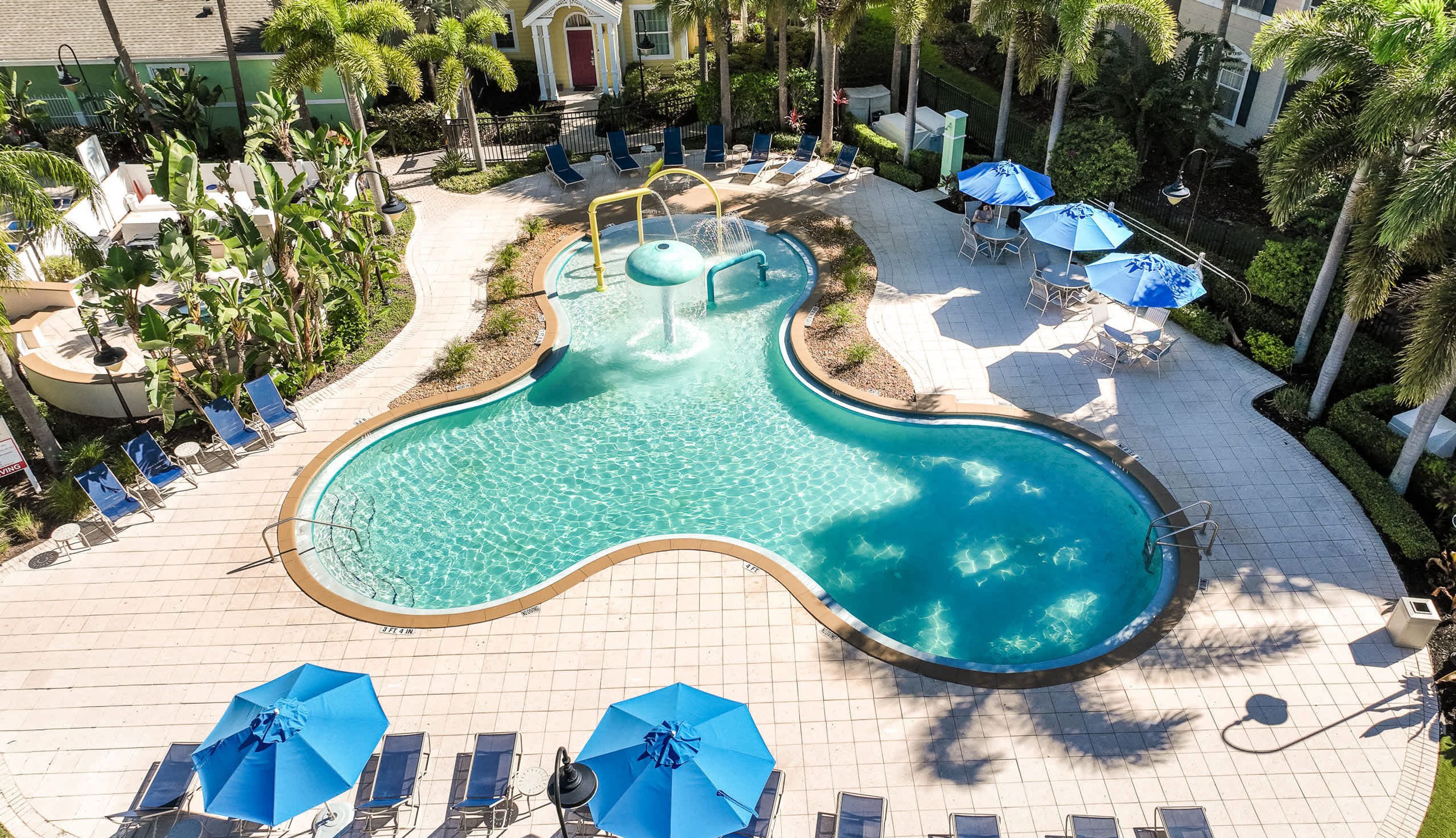 A pool with a unique shape surrounded by lounge chairs and umbrellas, set in a landscaped area with palm trees and a building in the background.