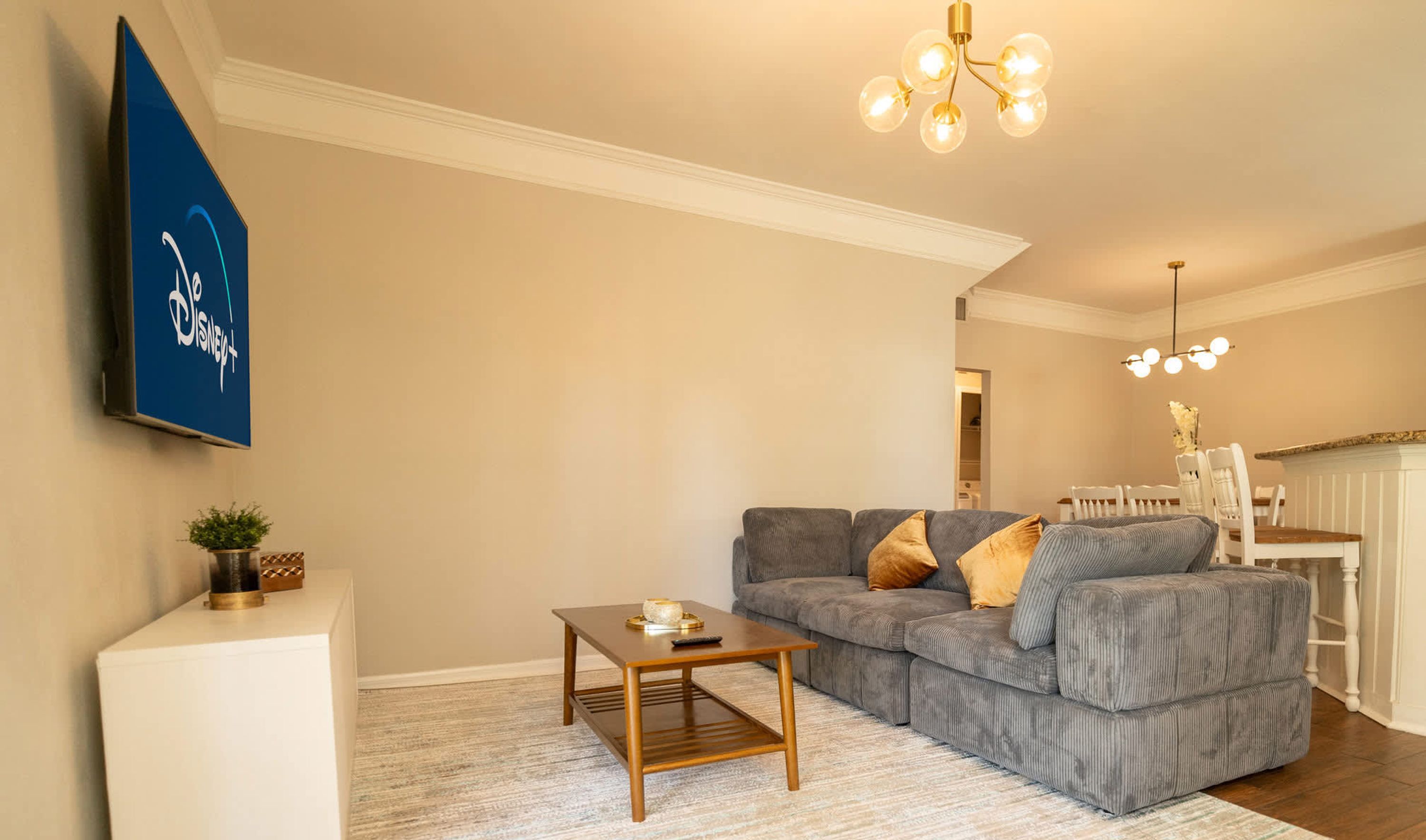 Ein Wohnzimmer mit grauem Sofa, einem Holztisch, einem an der Wand befestigten Fernseher und einer Kristallleuchter.