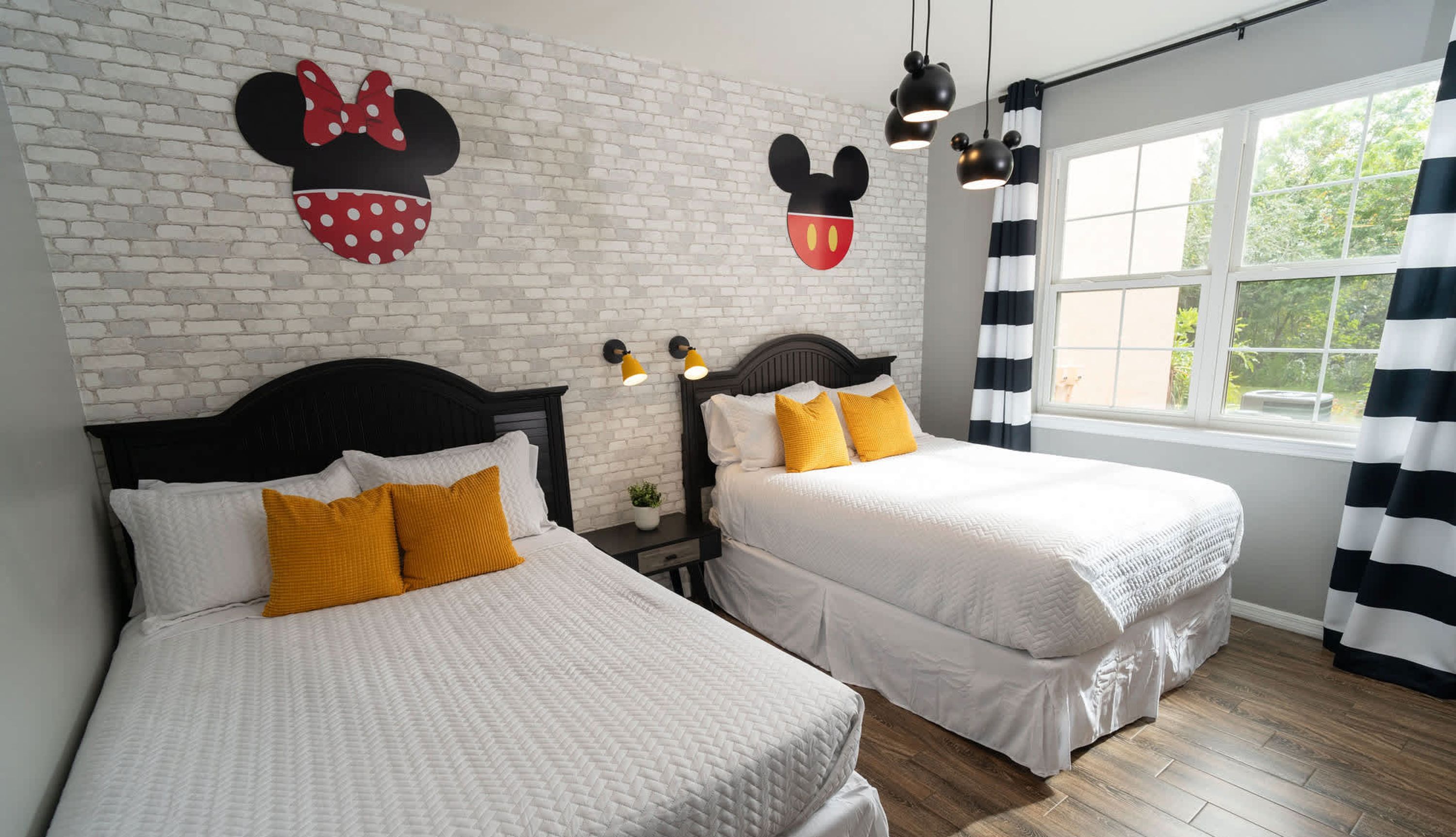 Habitación de hotel con dos camas, ropa de cama blanca, pared de ladrillo, decoración de Mickey Mouse y una ventana con cortinas.