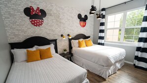 A hotel room with two beds, white bedding, a brick wall, Mickey Mouse decorations, and a window with curtains. - Magical Themed 3BR Townhouse Near the Parks (Kissimmee)