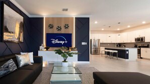 A modern living room with a dark-colored sofa, a glass coffee table, and a flat-screen TV displaying the Disney+ logo. - Luxury 6BR Villa w Pool Jacuzzi Fun in Orlando (Kissimmee)