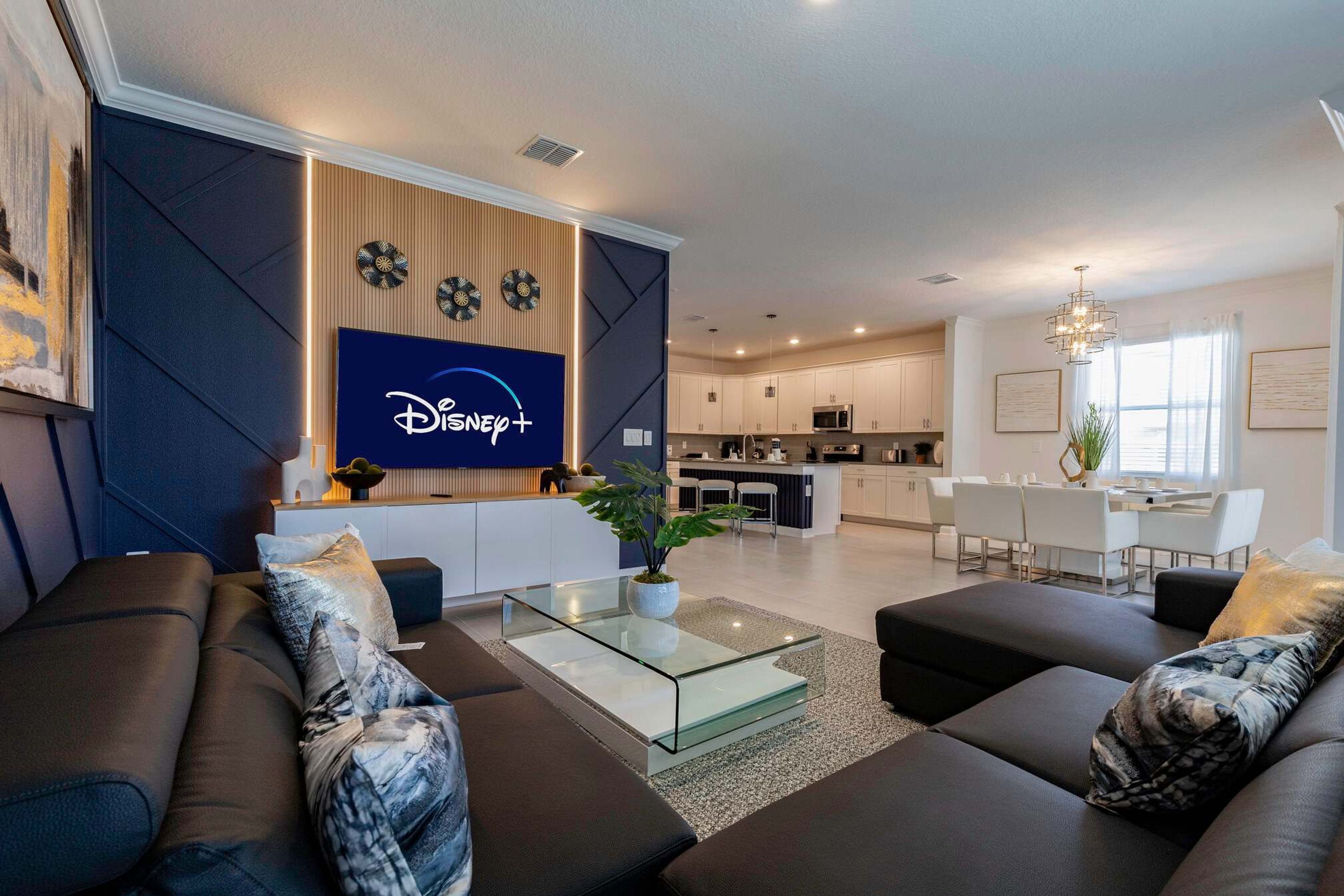 A modern living room with a glass coffee table, dark sofas, and a Disney+ logo on the wall.