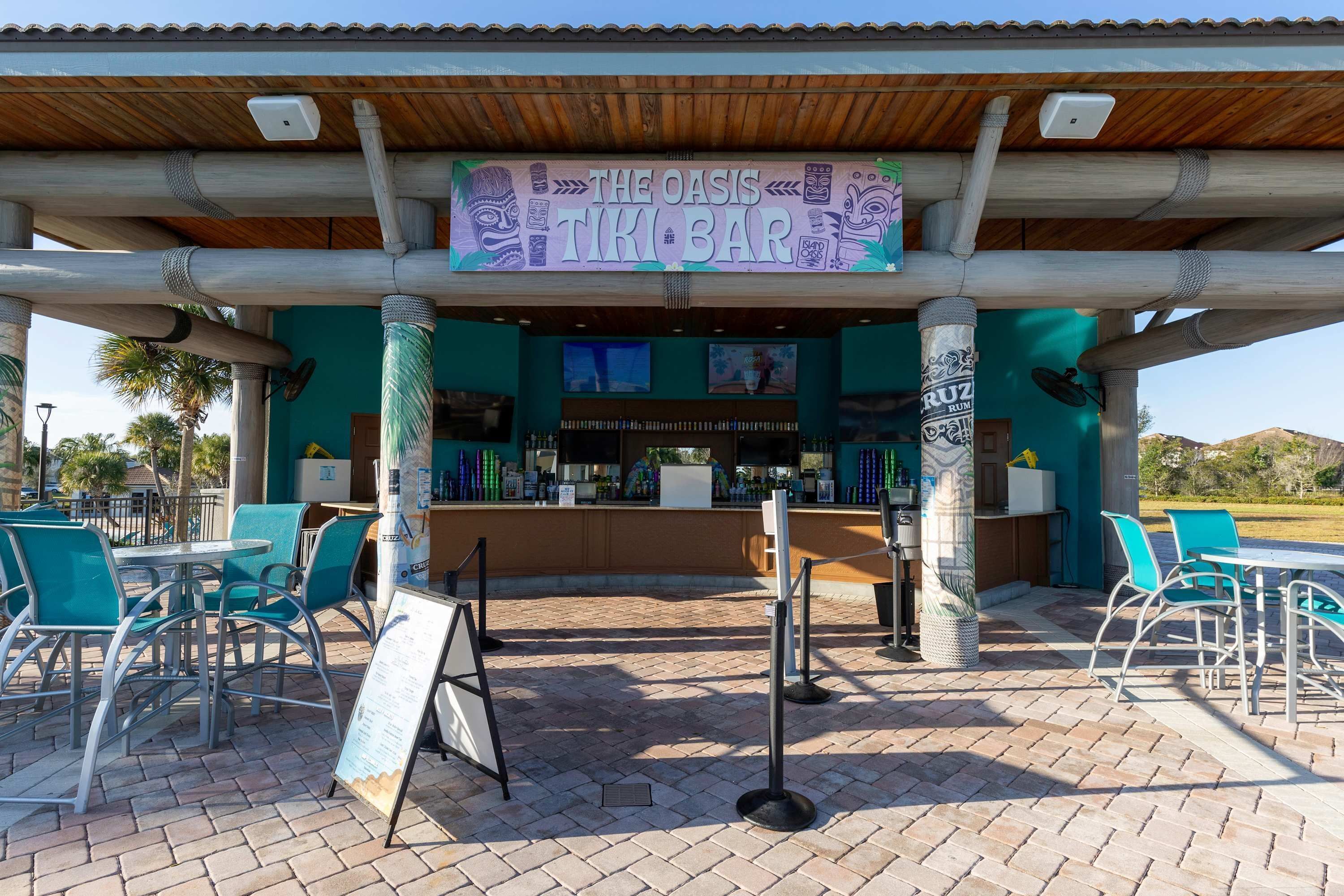 An outdoor bar area with a sign reading "The Oasis Tiki Bar," featuring a counter with a menu and a television screen. There are several tables and chairs arranged for seating.