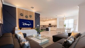 A modern living room with a glass coffee table, dark sofas, and a kitchen in the background. - Luxury 6BR Villa w Pool Jacuzzi Fun in Orlando (Kissimmee)
