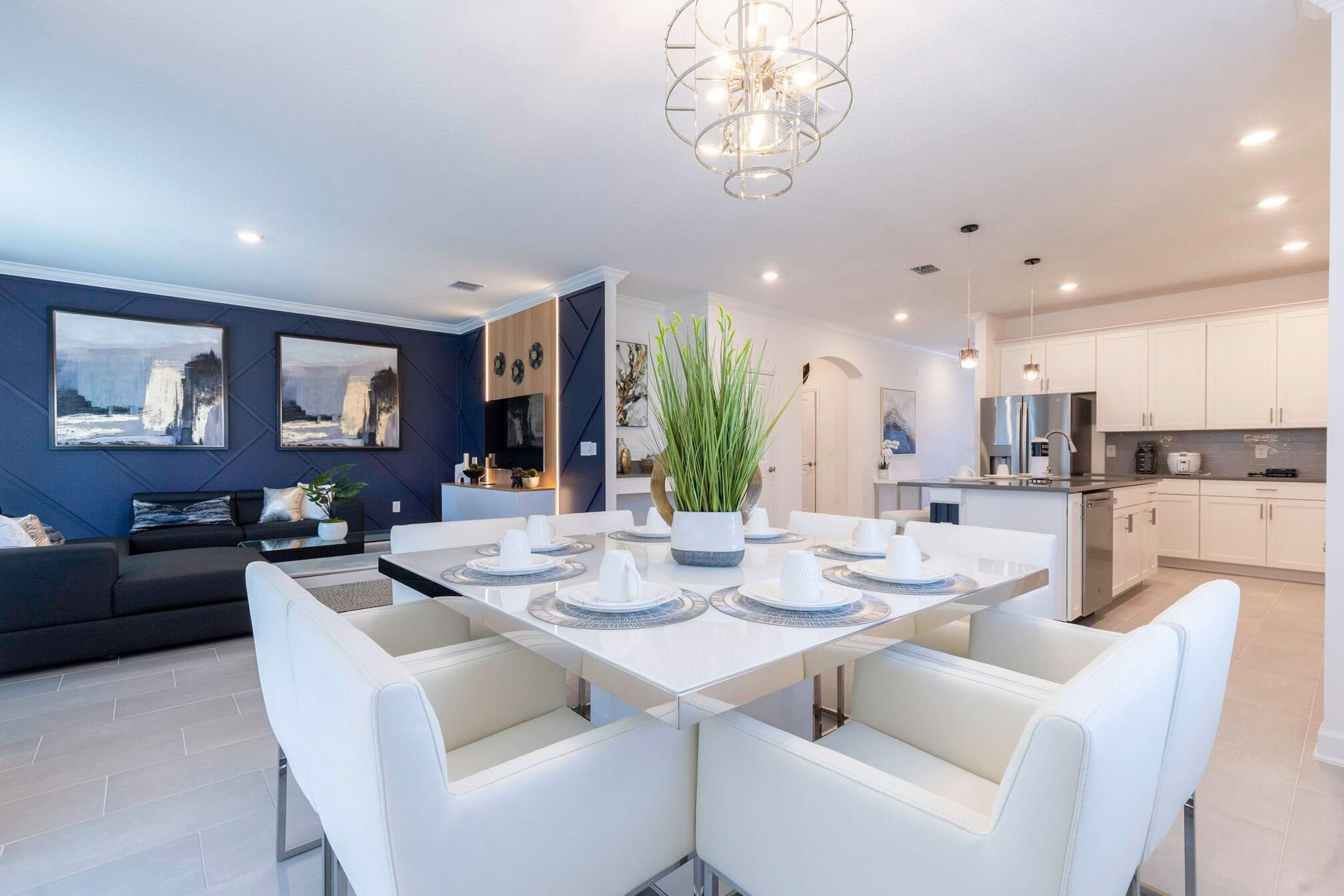 A modern dining area with a white table set for six, surrounded by white chairs. There is a large potted plant in the center.