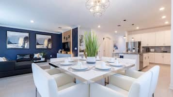 A modern dining area with a white table set for six, surrounded by white chairs. There is a large potted plant in the center.