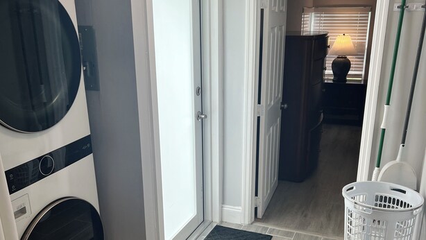 Un cuarto de lavandería con lavadora y secadora de carga frontal, una puerta que da a otra habitación, una cesta de ropa y un felpudo.