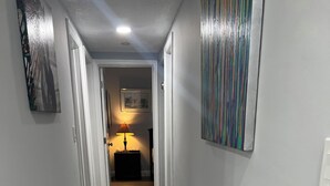 Un pasillo con piso de baldosas grises, paredes blancas y una puerta al final que da a una habitación con una lámpara y un cuadro en la pared.