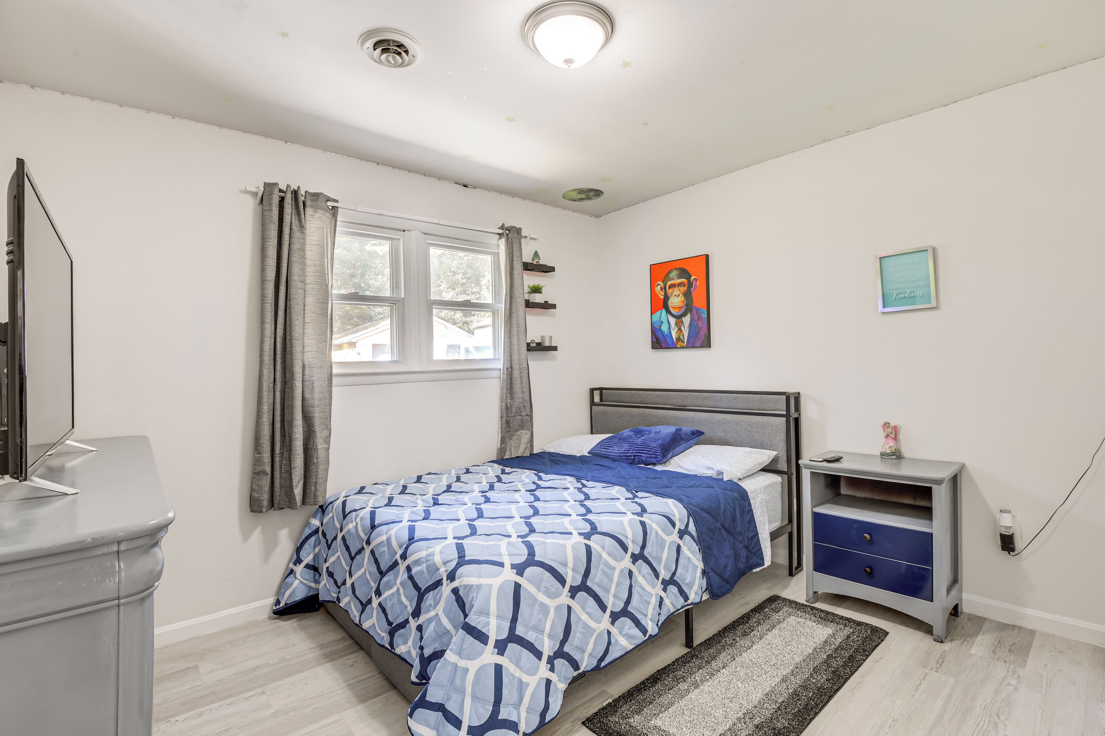 A bedroom with a bed, a nightstand, a dresser, a window with curtains, a painting on the wall, and a rug on the floor.