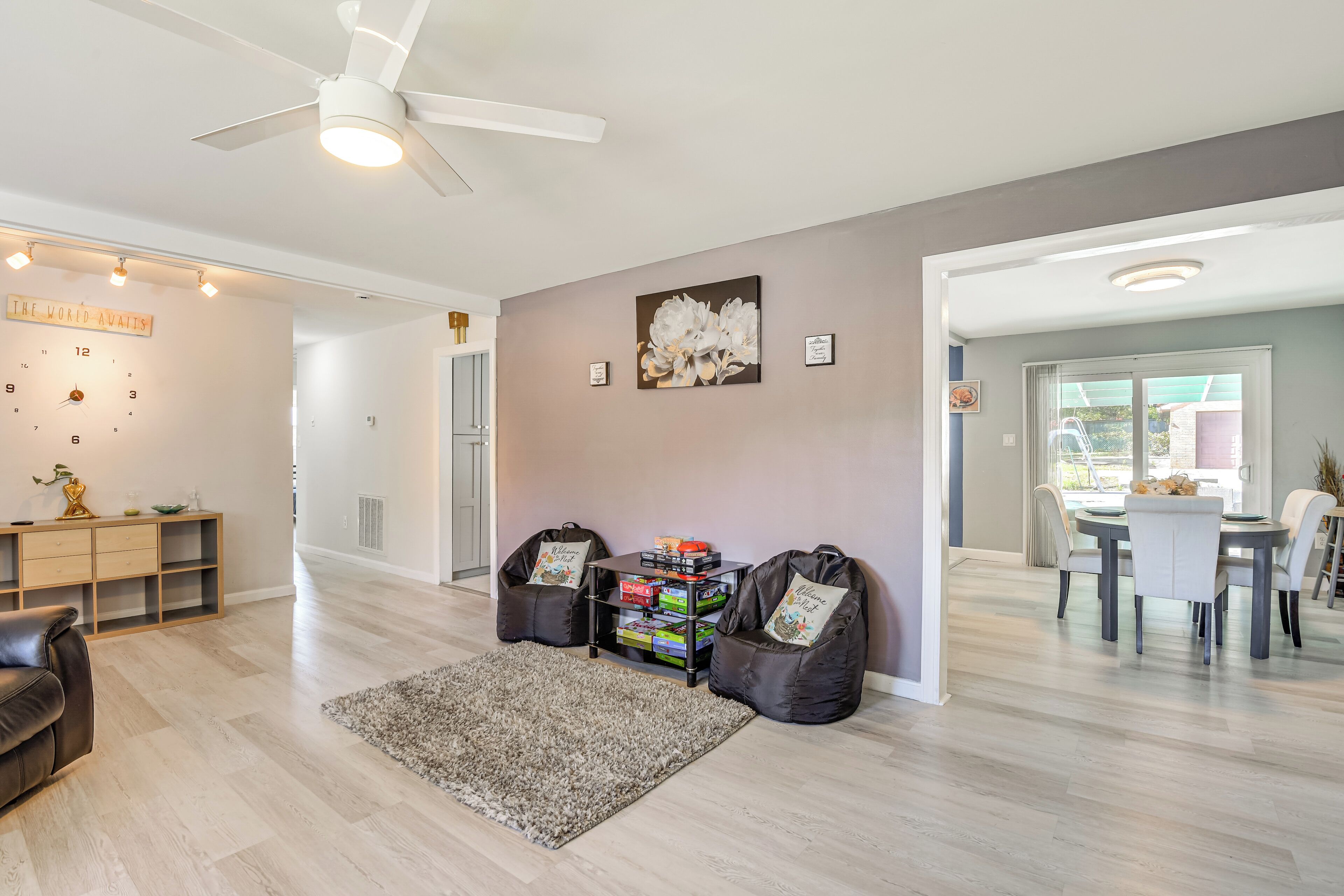 A spacious living room with a modern design, featuring a large ceiling fan, a wall clock, a bookshelf, and a dining area with a table and chairs.