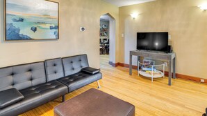 A living room with a black leather sofa, a brown ottoman, a flat-screen TV on a stand, and a painting on the wall. - 20 Min to Highmark Stadium: Yard & New Deck (Buffalo)