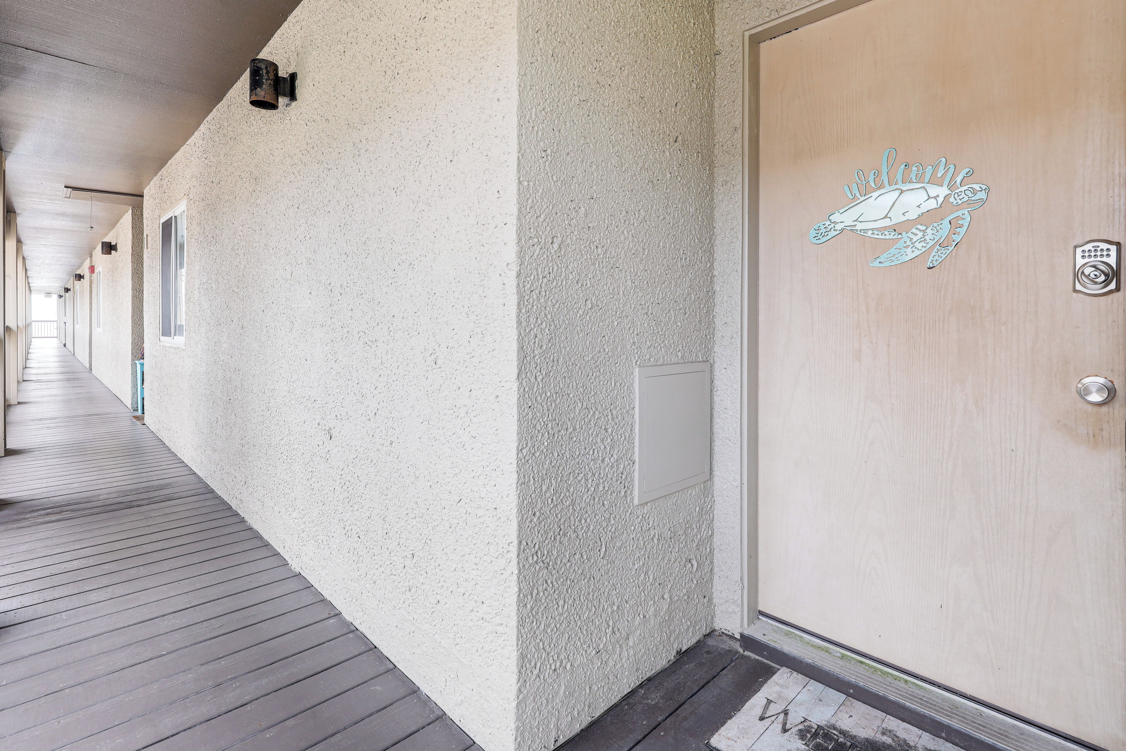 Un pasillo con piso de madera, pared texturizada y una puerta con letrero de 'welcome'.