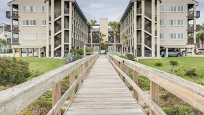 Een houten looppad leidt naar een modern appartementencomplex met meerdere balkons en een centrale binnenplaats.