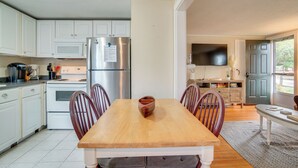 A modern kitchen with white cabinets, a stainless steel refrigerator, a wooden dining table with chairs, and a TV mounted on the wall. - Walk to Beaches & Dining: Classic Cape Cod Cottage (Dennis Port)