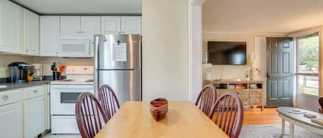 A modern kitchen with white cabinets, a stainless steel refrigerator, a wooden dining table with chairs, and a TV mounted on the wall.