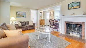 A living room with a fireplace, a dining area, and a painting on the wall. - Walk to Beaches & Dining: Classic Cape Cod Cottage (Dennis Port)