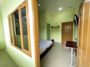 Una habitación con cama, una puerta de madera, una mesita pequeña, un cesto de basura y un televisor instalado en la pared.