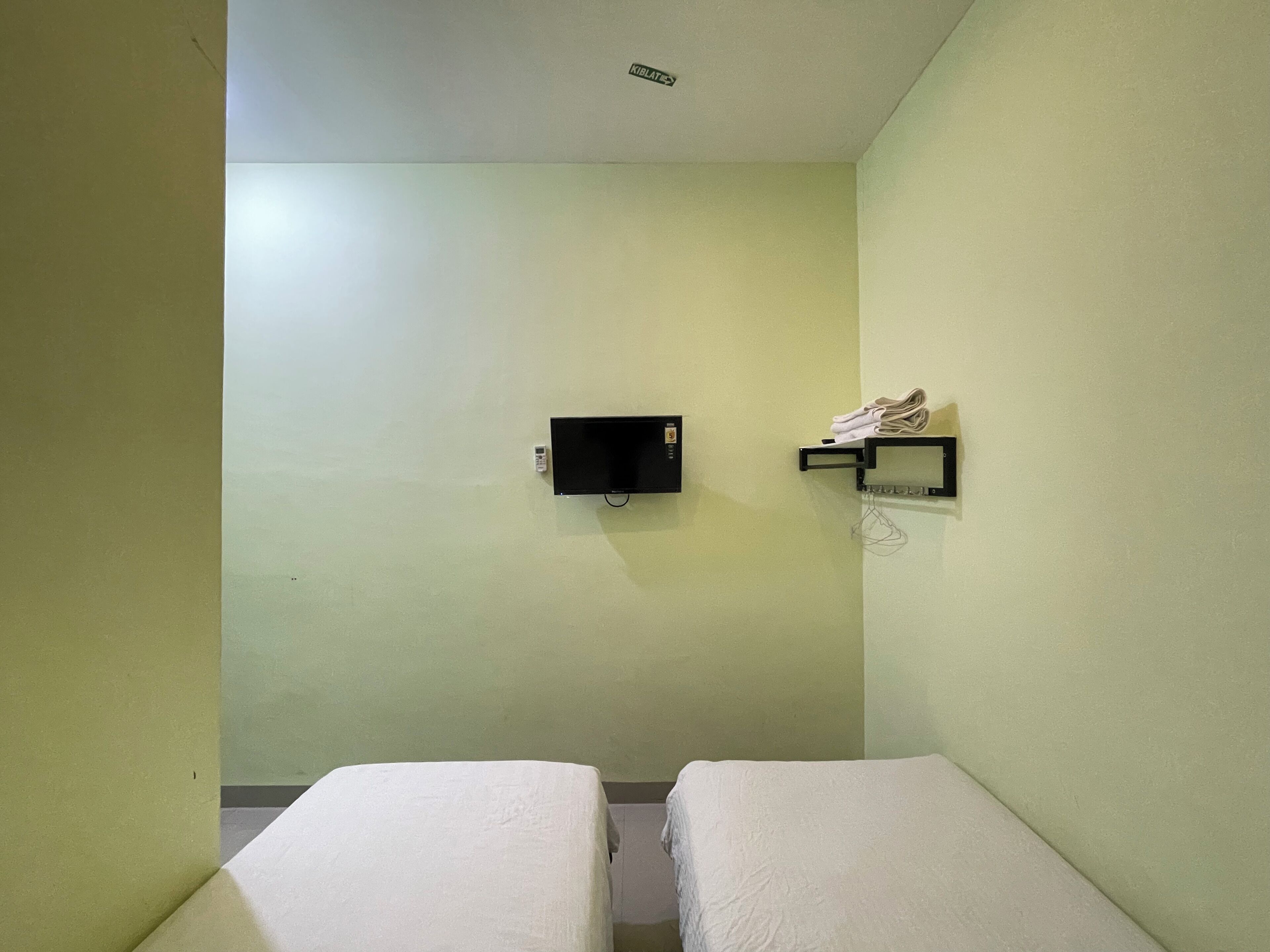A small, single-room hotel room with two beds, a TV mounted on the wall, and a shelf with a pair of shoes.