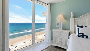 Una habitación con un amplio ventanal con vista a la playa, una cama con ropa de cama blanca, una mesita de noche con lámpara y una pared con un cuadro enmarcado.