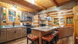 Une cuisine rustique aménagée dans une cabane en rondins, avec des murs en bois, un mur d’accent en pierre, un grand îlot central doté de tabourets et une cheminée en pierre.