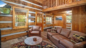 L’intérieur d’une cabane en rondins, avec un plancher en bois, un canapé en cuir, un fauteuil en cuir, une table basse ronde et un grand miroir.
